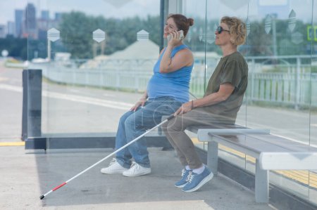 Eine ältere blinde Frau und eine schwangere Frau sitzen an einer Bushaltestelle und warten auf den Bus