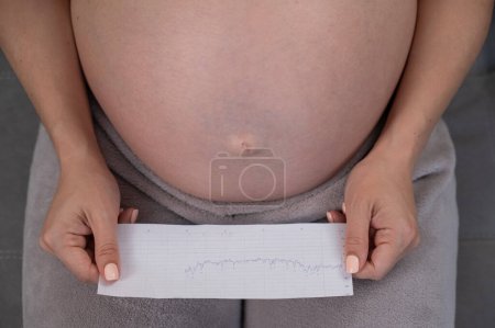 A pregnant woman looks at the results of cardiotocography while sitting on the couch. Close up of the belly
