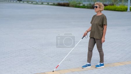 An elderly blind woman walks with a cane along a tactile tile