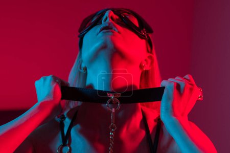 Foto de Mujer rubia en máscara de cuero ponerse el cuello en el dormitorio con luz de neón azul rojo - Imagen libre de derechos