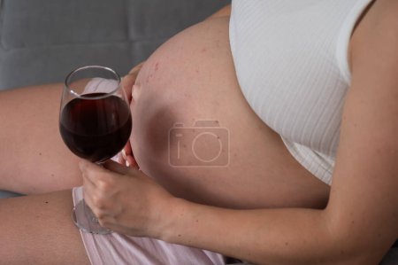 Großaufnahme des Bauches einer schwangeren Frau, die auf dem Sofa sitzt und ein Glas Rotwein hält. Hautausschlag
