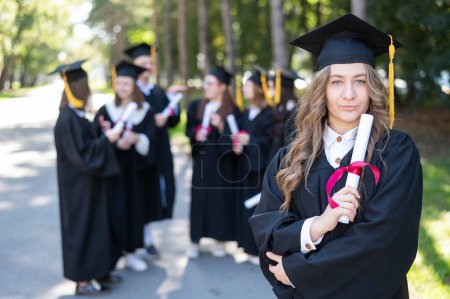 Téléchargez les photos : Groupe d'étudiants heureux en robes de remise des diplômes à l'extérieur. Une jeune fille avec un diplôme dans les mains au premier plan - en image libre de droit