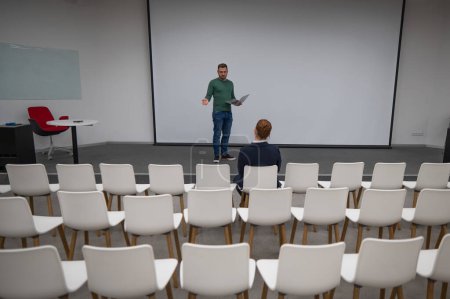 A red-haired Caucasian businesswoman sits in the front row of an empty conference room. Bearded man giving a lecture