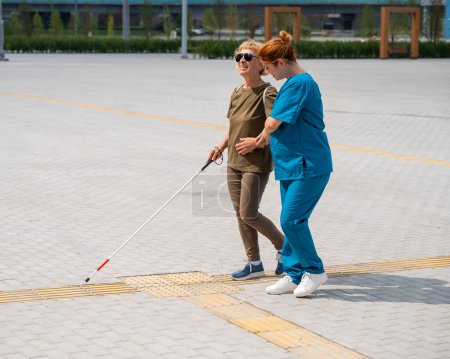A nurse accompanies an elderly blind woman on a walk