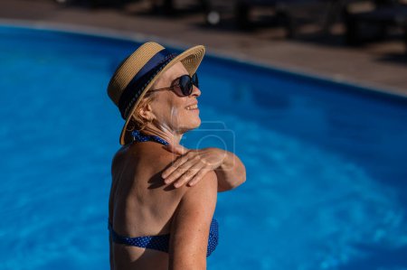 Photo for Portrait of an old woman in a straw hat, sunglasses and a swimsuit applying sunscreen to her skin while relaxing by the pool - Royalty Free Image