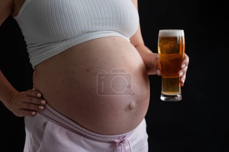 Großaufnahme des Bauches einer schwangeren Frau, die ein Glas Bier auf schwarzem Hintergrund hält. Hautausschlag