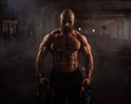 Musculoso hombre calvo sin camisa sosteniendo placas de peso. Un culturista muestra su forma en el gimnasio