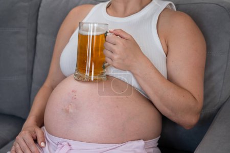 Eine gesichtslose Schwangere sitzt auf dem Sofa und hält sich ein Glas Bier auf den Bauch. Hautausschlag