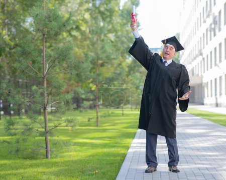 A serious old man in a graduate robe pulls his hand with a diploma up outdoors