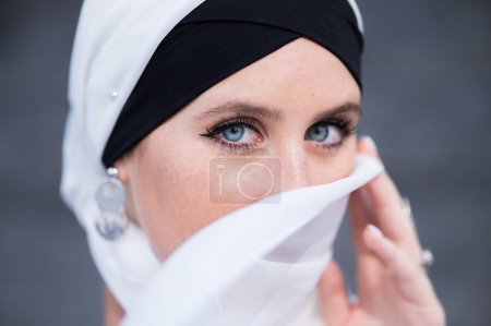 Retrato de una joven mujer de ojos azules en un hiyab contra una pared de ladrillo gris. Mujer musulmana con bufanda blanca cubriéndose la cara