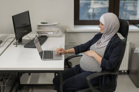 Schwangere Kaukasierin im Hidschab arbeitet am Computer im Büro