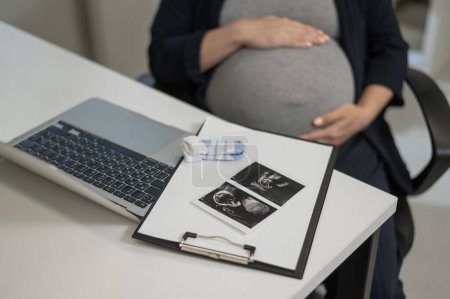 A pregnant woman works on a laptop in the office and looks at a photo from an ultrasound scan of the fetus. Belly close-up