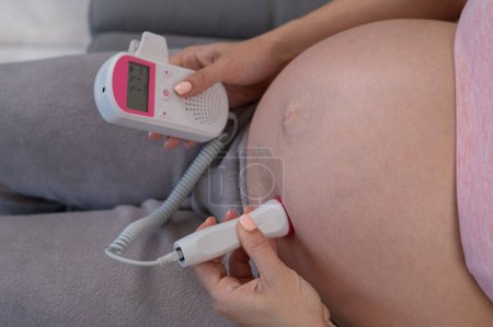 Téléchargez les photos : Gros plan d'un ventre de femme enceinte à l'aide d'un doppler à domicile. Lectures erronées. Battements de cœur des mères - en image libre de droit