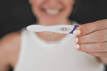 Téléchargez les photos : Portrait flou d'une femme caucasienne heureuse tenant un test de grossesse express positif sur fond noir - en image libre de droit