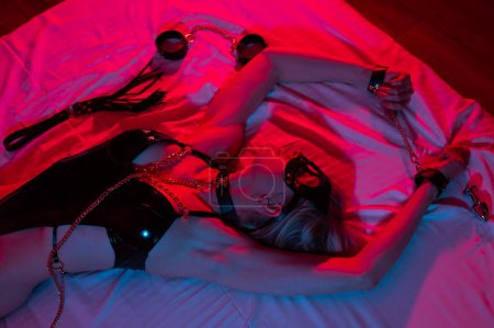 Foto de Mujer rubia sexy con una máscara de cuero y esposas y un collar se encuentra en el dormitorio en una luz de neón rojo-azul. Juegos de rol - Imagen libre de derechos