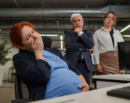 Foto de La mujer embarazada bosteza en el trabajo. Colegas miran desaprobadamente - Imagen libre de derechos
