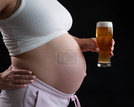 Großaufnahme des Bauches einer schwangeren Frau, die ein Glas Bier auf schwarzem Hintergrund hält. Hautausschlag