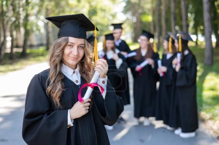 Téléchargez les photos : Groupe d'étudiants heureux en robes de remise des diplômes à l'extérieur. Une jeune fille avec un diplôme dans les mains au premier plan - en image libre de droit