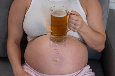 Téléchargez les photos : Une femme enceinte sans visage est assise sur le canapé et tient un verre de bière sur le ventre. Éruption cutanée - en image libre de droit