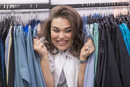 Téléchargez les photos : Une grosse femme dans un magasin de plus de taille jette un coup d'oeil derrière des rayonnages de vêtements - en image libre de droit