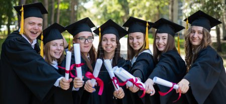 Téléchargez les photos : Rangée de jeunes gens heureux en robes de remise des diplômes titulaires de diplômes en plein air - en image libre de droit