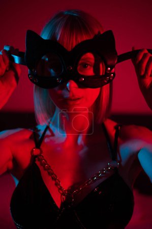 Foto de Mujer rubia que se pone la máscara de cuero en el dormitorio en luz de neón azul rojo - Imagen libre de derechos