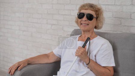 An elderly blind woman wearing sunglasses and with a tactile cane sits on the sofa