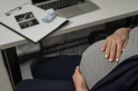 Una mujer embarazada trabaja en una computadora portátil en la oficina y mira una foto de una ecografía del feto. Primer plano del vientre