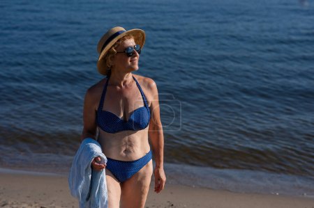 Photo for An old woman in a straw hat, sunglasses and a swimsuit is resting on the beach - Royalty Free Image