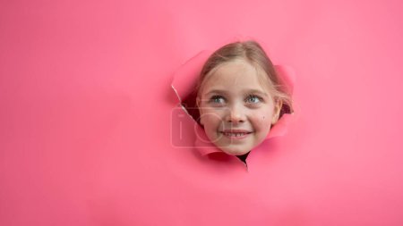Niedliche kaukasische Mädchen guckt aus einem Loch in einem Papier rosa Hintergrund