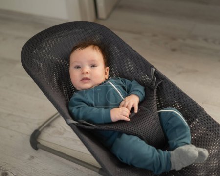 Niedliches Neugeborenes in blauen Overalls sitzt in einer Babyliege