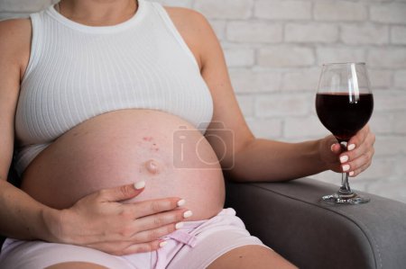 Close-up of the belly of a pregnant woman holding a glass of red wine while sitting on the sofa. Skin rash
