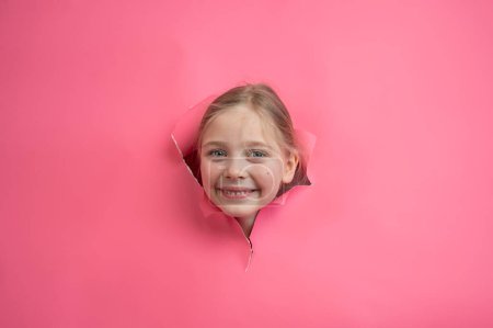 Cute Caucasian girl peeks out of a hole in a paper pink background