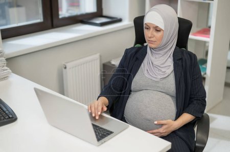 Schwangere Kaukasierin im Hidschab arbeitet am Computer im Büro