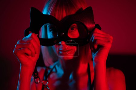 Foto de Mujer rubia que se pone la máscara de cuero en el dormitorio en luz de neón azul rojo - Imagen libre de derechos