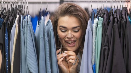 Téléchargez les photos : Une grosse femme dans un magasin de plus de taille jette un coup d'oeil derrière des rayonnages de vêtements - en image libre de droit