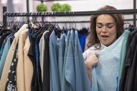 Téléchargez les photos : Portrait d'une grosse femme dans un magasin de taille plus à travers des cintres avec des vêtements - en image libre de droit