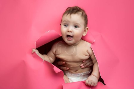 Cute Caucasian newborn baby boy peeks out of a hole in a paper pink background