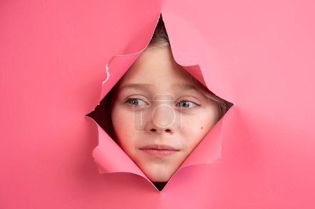Cute Caucasian girl peeks out of a hole in a paper pink background