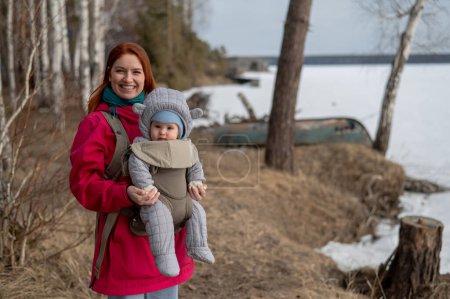 Caucasian red-haired woman walks with her son in an ergo backpack in nature in winter