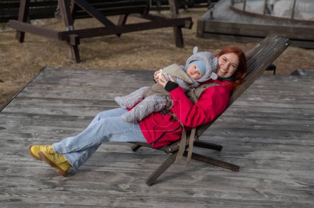 Caucasian woman with her son in an ergo backpack sitting in a wooden deck chair