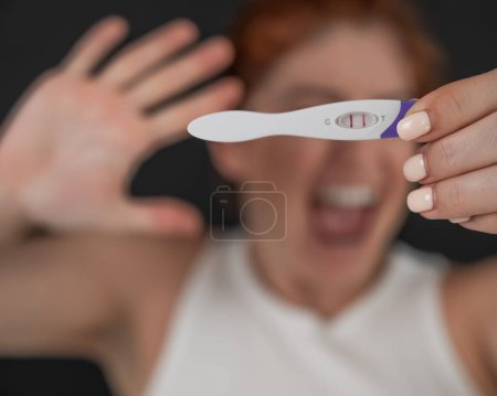 Téléchargez les photos : Portrait flou d'une femme caucasienne bouleversée tenant un test de grossesse rapide positif sur fond noir - en image libre de droit