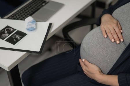 Una mujer embarazada trabaja en una computadora portátil en la oficina y mira una foto de una ecografía del feto. Primer plano del vientre