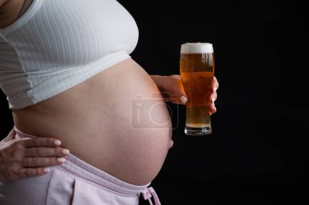 Close-up of the belly of a pregnant woman holding a glass of beer on a black background. Skin rash