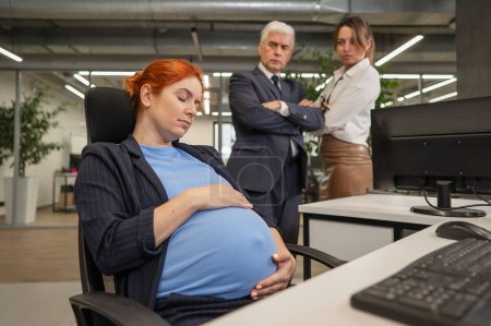 A pregnant woman sleeps at her workplace. Colleagues look disapprovingly