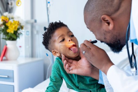 African american male doctor examining mouth of boy patient with penlight in hospital. Hospital, medical and healthcare services.