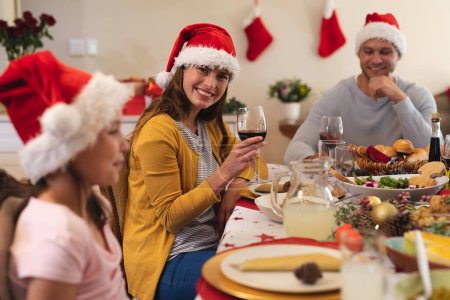 Portrait of caucasian woman sitting at table for dinner, wearing santa hat, holding glass of wine looking at camera and smiling, with her family in the background. christmas celebration.