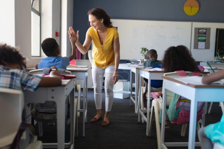 Happy caucasian young female teacher giving high five to african american boy sitting at desk. unaltered, education, childhood, teaching, learning and school concept.