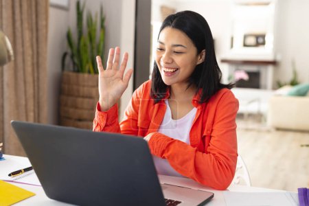 Image of happy biracial woman sitting at desk waving during video call on laptop at home, copy space. Working from home, technology, communication and lifestyle concept. Poster 654902072