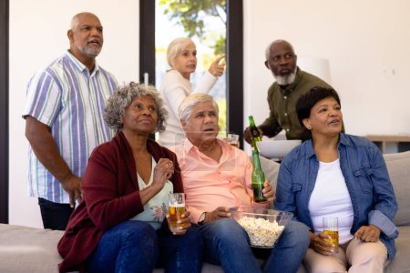 Multiracial senior friends with beer and popcorn watching soccer match in nursing home. Food, sport, curiosity, alcohol, unaltered, togetherness, support, assisted living and retirement concept.